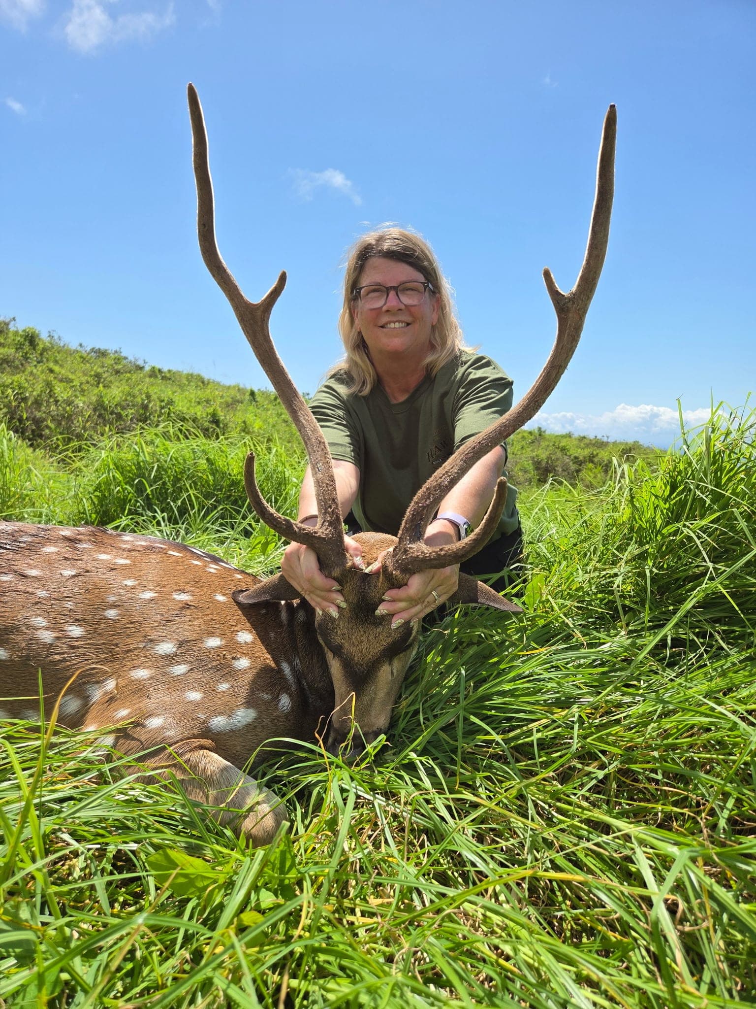A smiling hunter kneels behind a tall-antlered axis buck in deep green grass under a blue sky