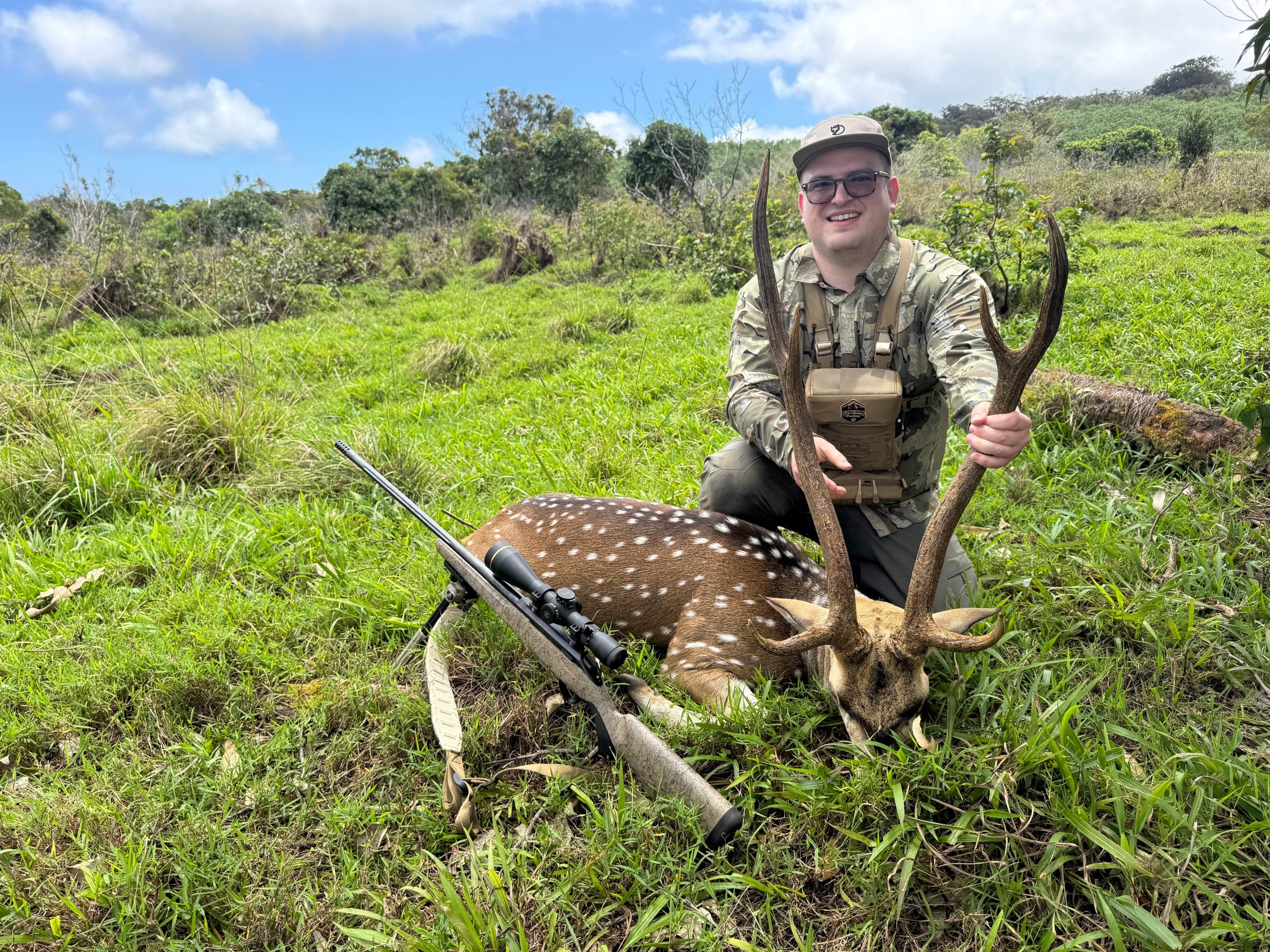 A hunter in camouflage kneels behind a spotted axis buck in bright green pasture under a blue Maui sky, rifle resting in the grass