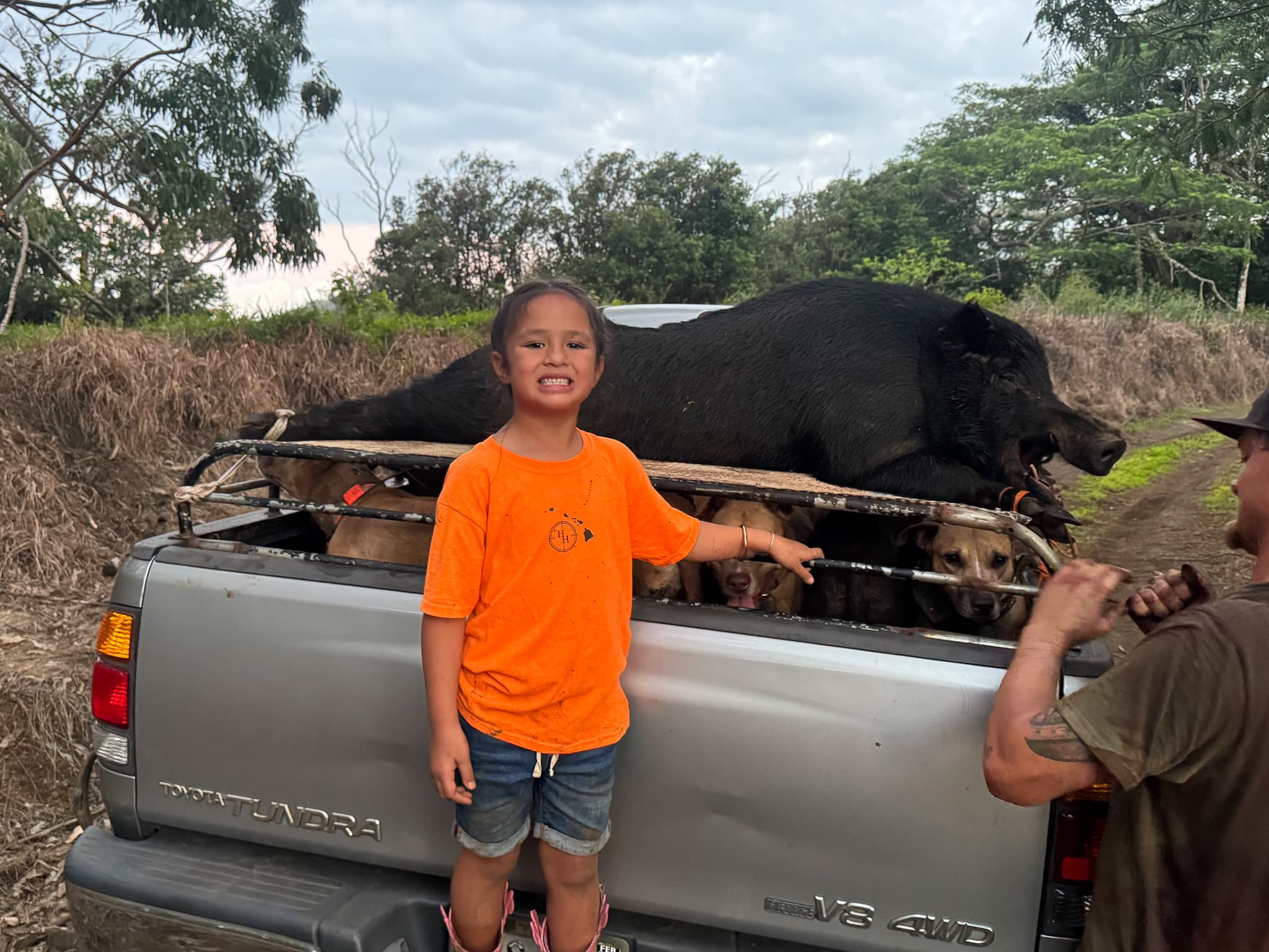 A young child stands proudly at the tailgate of a truck loaded with feral pigs after a family hunt