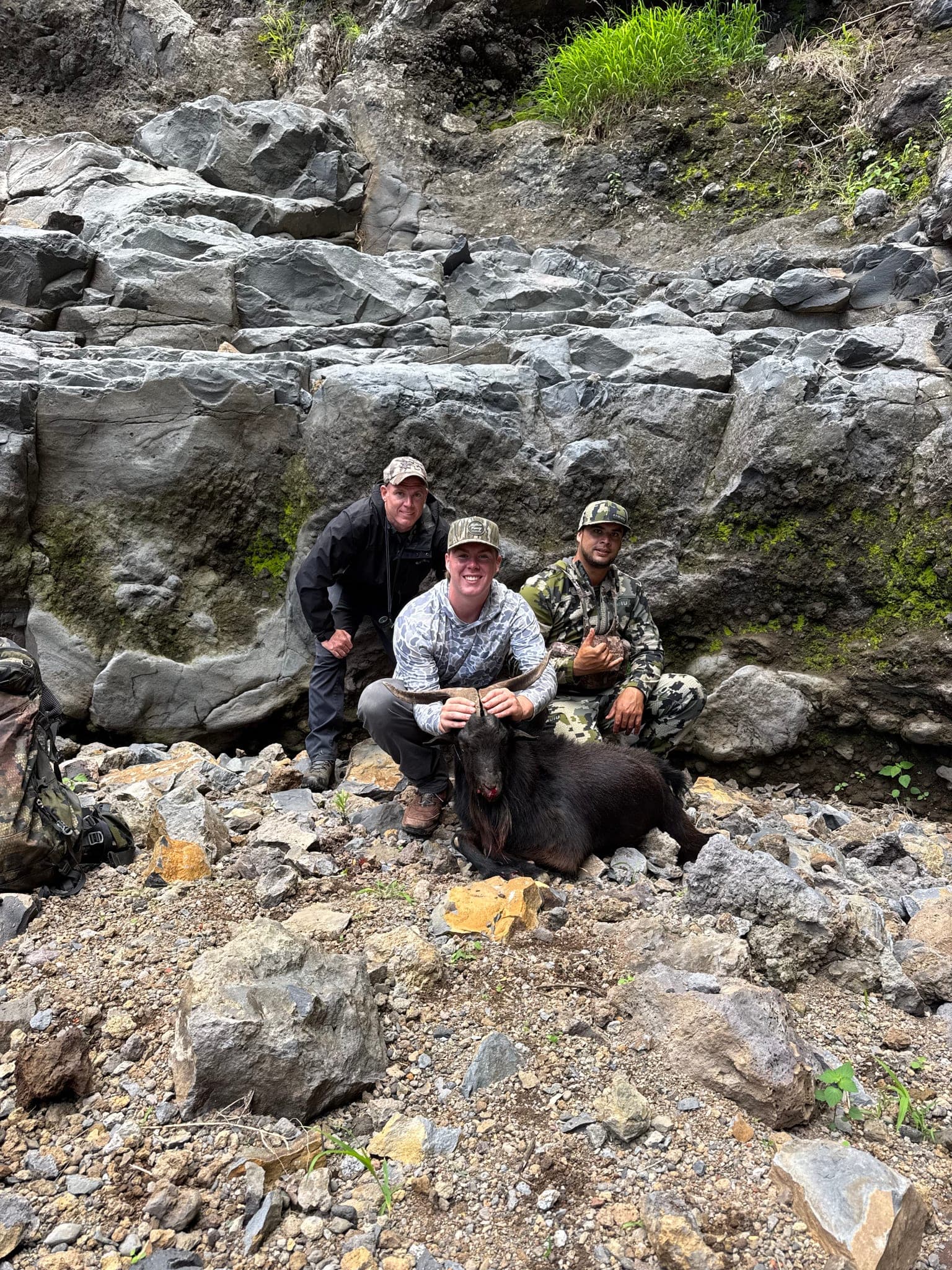 Three hunters kneel with a black mountain goat in a steep, rocky lava gulch