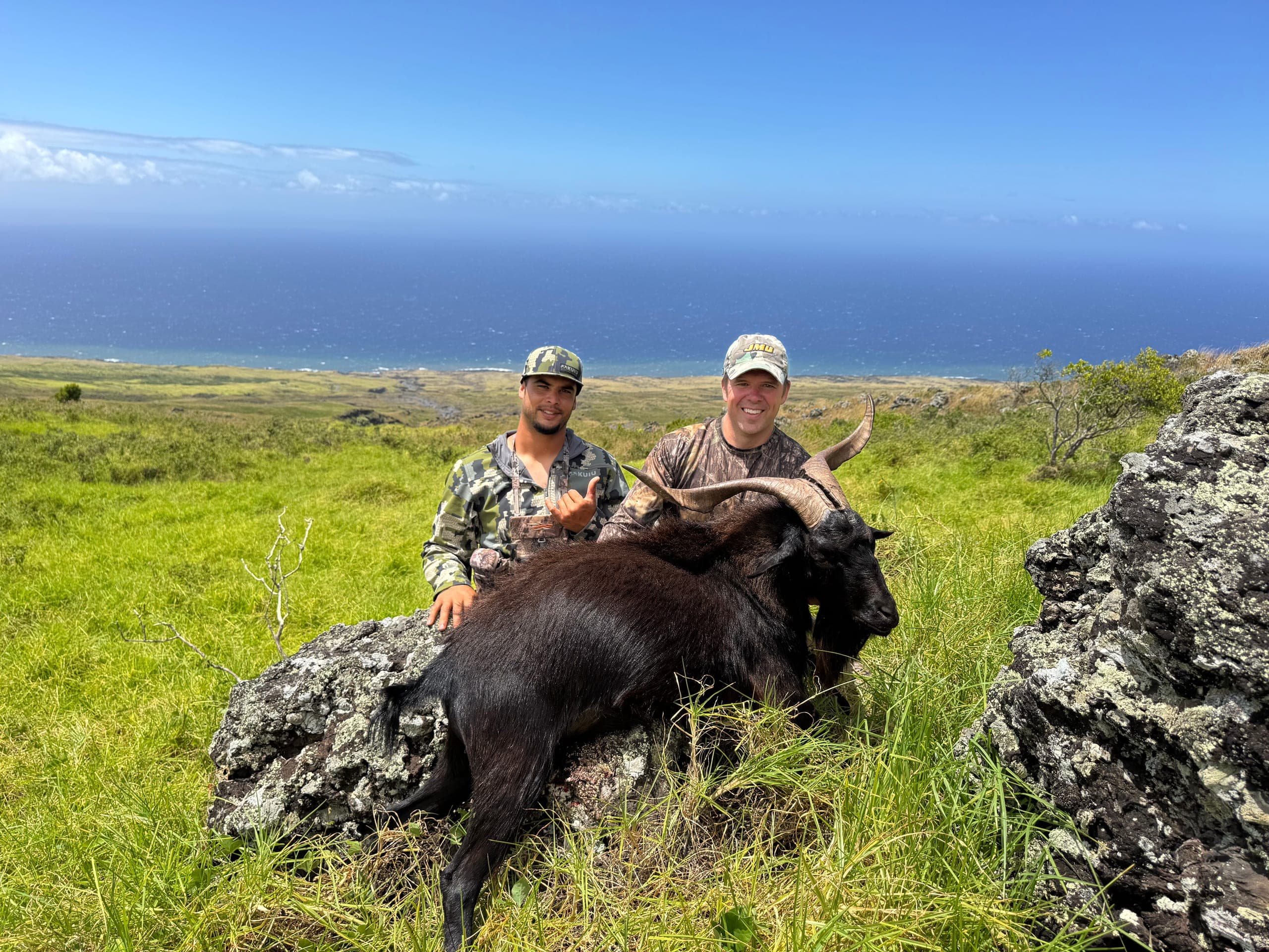 Two hunters kneel with a black mountain goat on a grassy lava bluff high above the deep blue ocean on Maui